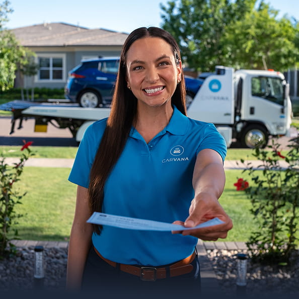 A Carvana employee at a customer’s house, delivering a check, with the customer’s car on a Carvana hauler in the background