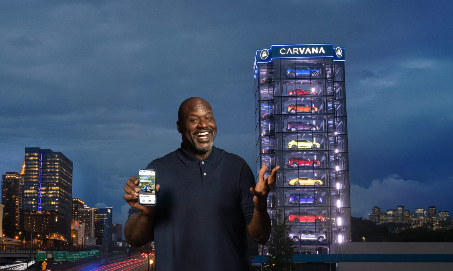 Shaquille O'Neal holding a phone displaying the Carvana app, standing near Carvana's car vending machine tower