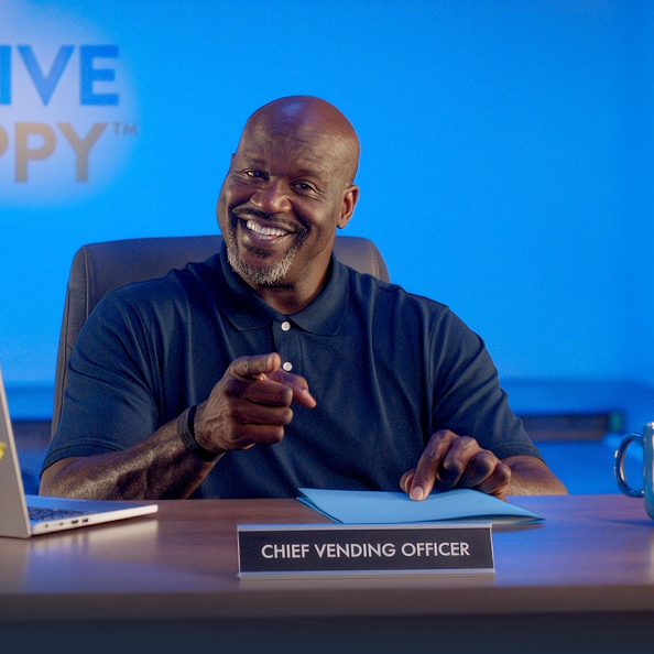 Shaquille O’Neal smiling and pointing at the camera from a desk with a miniature Carvana car vending machine
